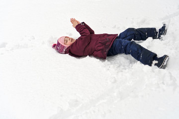 Little cute girl in winter outdoors. She lies in snow and makes snow angel. Girl rejoices and fools
