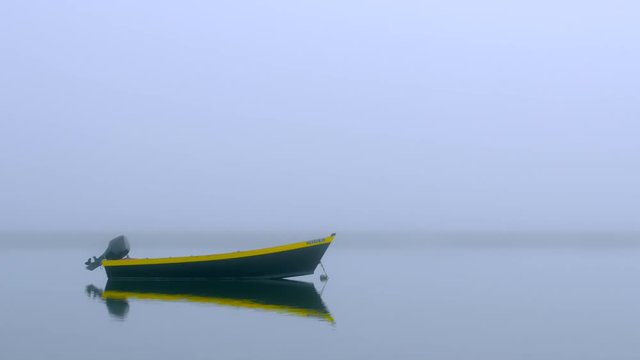 Black and yellow dinghy fishing boat at anchor in a quiet foggy cove with calm water and dune grass.