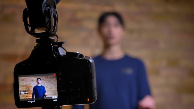 Closeup focused shoot of camera recording young korean male videoblogger talking and gesturing refocusing on the speaker