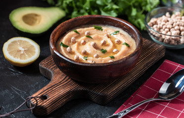 Hummus in to a brown wooden plate. On the black stone table are vegetables, greens, triangular pieces of pita. Top view. 