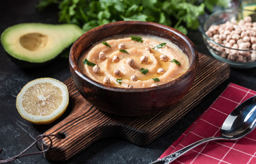 Hummus in to a brown wooden plate. On the black stone table are vegetables, greens, triangular pieces of pita. Top view. 