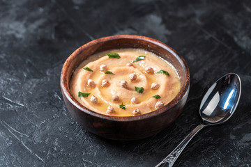 Hummus in to a brown wooden plate. On the black stone table are vegetables, greens, triangular pieces of pita. Top view. 