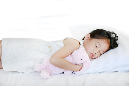Sleeping little Asian kid girl lying on bed with hugging doll.