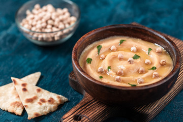 Hummus in to a brown wooden plate. On the blue stone table are vegetables, greens, triangular pieces of pita.