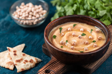 Hummus in to a brown wooden plate. On the blue stone table are vegetables, greens, triangular pieces of pita.