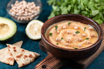 Hummus in to a brown wooden plate. On the blue stone table are vegetables, greens, triangular pieces of pita.