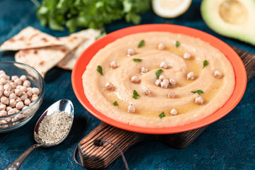 Hummus in the orange plate. On the blue stone table are vegetables, greens, triangular pieces of pita.