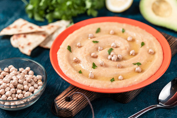 Hummus in the orange plate. On the blue stone table are vegetables, greens, triangular pieces of pita.
