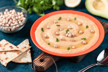 Hummus in the orange plate. On the blue stone table are vegetables, greens, triangular pieces of pita.