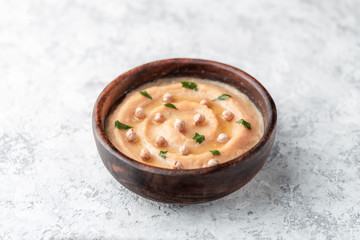 Hummus in the brown wooden plate. On the white table are vegetables, greens, triangular pieces of pita.