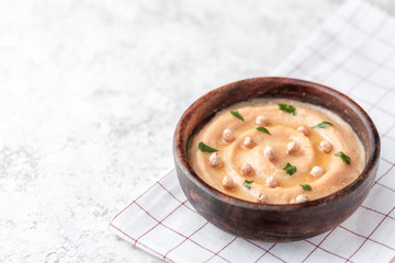 Hummus in the brown wooden plate. On the white table are vegetables, greens, triangular pieces of pita.