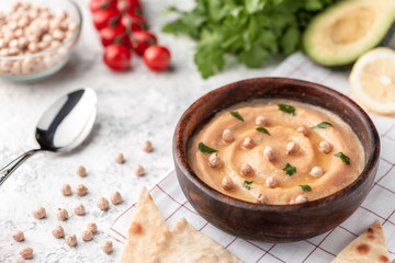 Hummus in the brown wooden plate. On the white table are vegetables, greens, triangular pieces of pita.