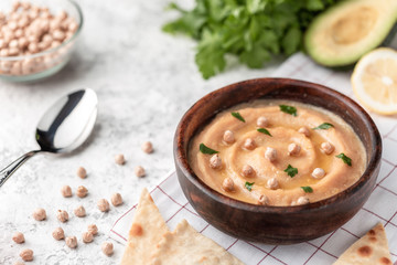 Hummus in the brown wooden plate. On the white table are vegetables, greens, triangular pieces of pita.