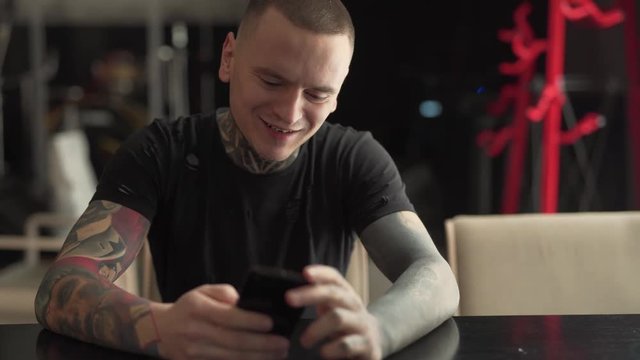 Young man in black t-shirt with tattoos on his neck and arms is sitting in a cafe indoors, looking at his phone, clicking, chewing a buble gum and smiling a lot. Man is looking like a rockstar.