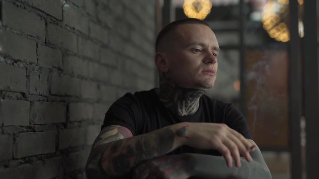 Young man with a many tattoos is sitting in a cafe indoors, smoking, chewing a gum and thinking. Man is sitting near brics wall and looking pensive and thoughtful.