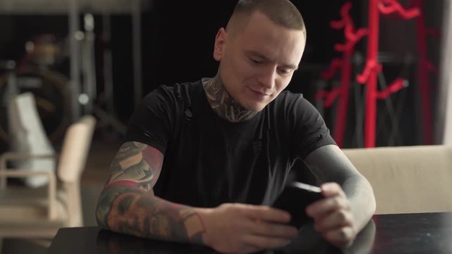 Young man with tattoos on his neck and upper limb in a dark t-shirt is sitting in a cafe indoors, looking at his phone, clicking and smiling. Man is looking absolutely delighted.