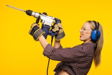 Beautiful young woman in goggles and headphones worth holding perforator drill isolated on yellow...