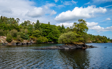Fototapeta na wymiar Rocky island with a couple of trees, in welsh lake. Background of blue sky and white puffy clouds