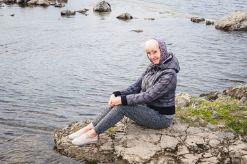 cute woman fifty years in a gray jacket and leggings sitting on a stone near the river