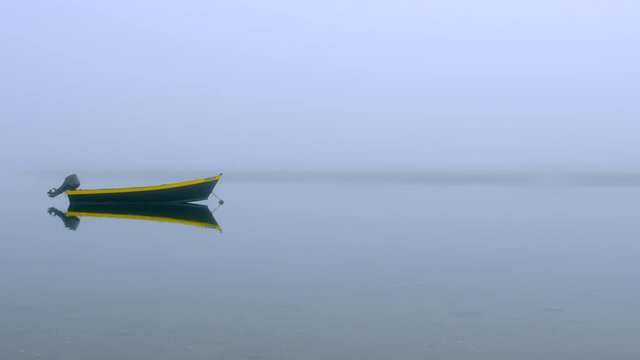 Black and yellow dinghy fishing boat at anchor in a quiet foggy cove with calm water and dune grass.