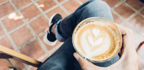 Young man in blue jeans and wearing black shoes sitting on chair holding cup of coffee latte art as relax time at coffee shop in the morning