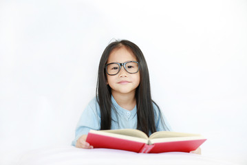 Smiling little Asian kid girl wearing glasses reading hardcover book lying on bed against white background.