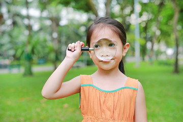 Cute little Asian child girl looking through magnifiying glass on at grass outdoors.