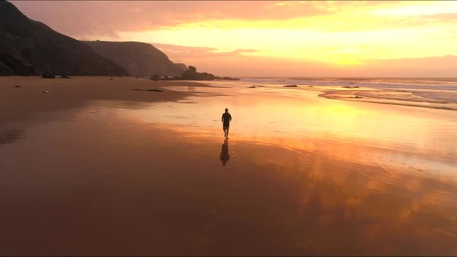 A man running along the beach while sunset and water refection the water at the red and orange cliffs and coastline. Aerial helicopter drone shot following