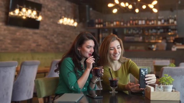 Two girls are sitting in a cafe indoors and talking in a video chat with their friend. Both woman are drinking a mulled wine. They are looking absolutely delighted and cheerful to see their friend.