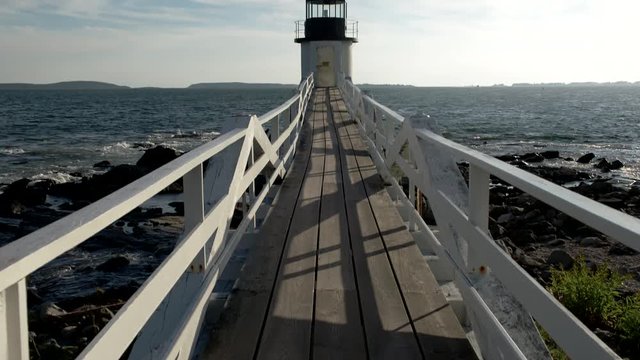 Maine coastline lighthouse with long wooden boardwalk leading out over the water. Camera tilts up from the wooden planks.