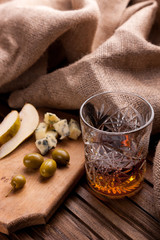 Rustic still life with whiskey and snacks. Crystal glass with a beautiful pattern on a wooden table. Snack for whiskey on crumpled paper olives, crackers, cheese, almonds, pear, apple.