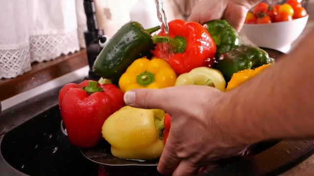 Male hands wash lots of bell peppers at the kitchen sink - holding a strainer under the tap water flowing, slow motion, camera slide