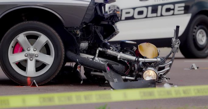 Damaged motorcycle is wedged under the bumper of a car after a fatal traffic accident.  Police car and evidence markers surround the crash scene where the motorcyclist was killed.