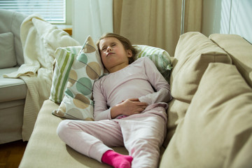 Child lying on sofa in the living room with stomach pain. Hands on belly. Little girl suffering