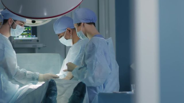 Tracking shot of concentrated surgical team wearing sanitary clothing, caps, masks, and gloves while operating on patient at hospital