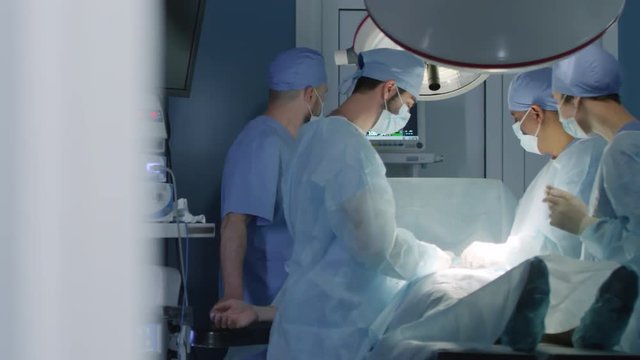 Tracking shot of four concentrated surgeons performing medical operation on patient at hospital