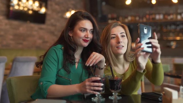 Two young women are talking in a video chat with their friend. Both woman are drinking a mulled wine. They are looking absolutely happy and satisfied to see their friend.