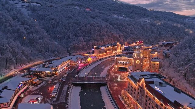 Aerial drone shot. Fly above Rosa Khutor ski resort in Sochi. evening illumination. Snow covered forest. Flying forward above river.