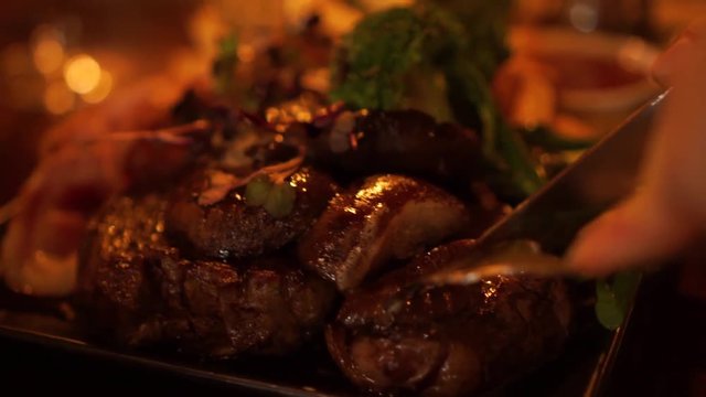 Cutting a steak on a plate with mushrooms, bacon, broccoli, salad, french fries and gravy at a luxury restaurant - CLOSE UP