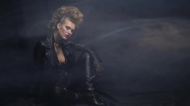 Rocker girl in black leather clothes, with crazy hairstyle, posing on the floor in the smoke