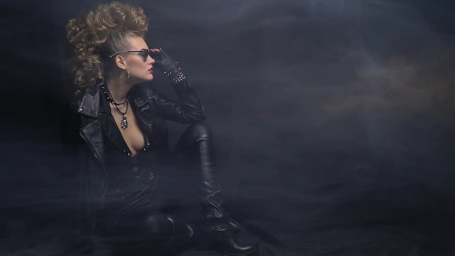 Rocker woman in leather jacket, posing on the floor in sunglasses, smoke and black background
