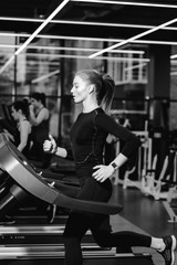 Black and white photo of the athletic girl dressed in a black sportswear running on the treadmill in the modern gym