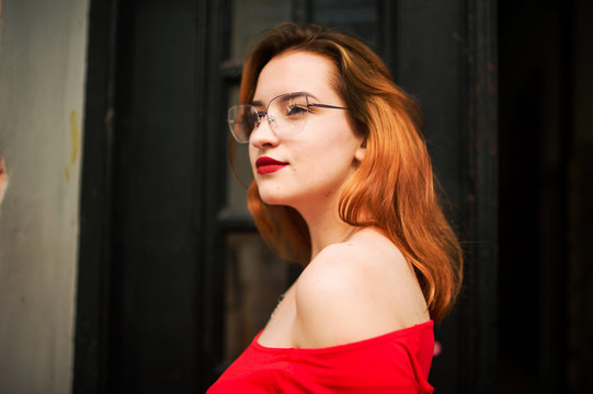 Attractive redhaired woman in eyeglasses, wear on red blouse posing at street against old wooden door.