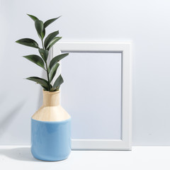 Mock up white frame and branch with green leaves in blue vase on book shelf or desk. Minimalistic concept.