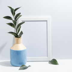 Mock up white frame and branch with green leaves in blue vase on book shelf or desk. Minimalistic concept.