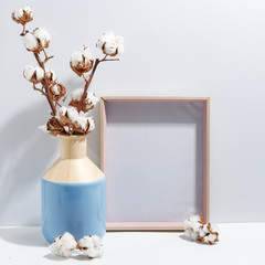 Mock up white frame and dry cotton twigs in blue vase on book shelf or desk. Minimalistic concept.