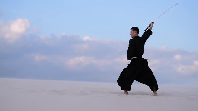 Concentrated man, in traditional Japanese clothes, with sword, katana, is training martial arts in desert during sunset - samurai on the blue cloudy sky background. 