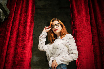 Attractive redhaired woman in eyeglasses, wear on white blouse posing at arch of open red curtains.