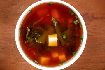 A closeup photo of a bowl of miso shiru soup with tofu, scallions, and wakame seaweed, shot from the top on a rustic wooden background