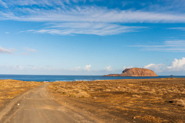 Empty sand road through an arid volcanic landscape leading towards the blue sea under a clear summer sky on a hot day. La Graciosa Island seascape, Canary, Spain.